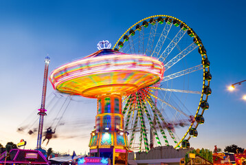 Funfair panorama wiht ferris wheel and carousels on big german funfair or Kirmes at blue hour twilight. Long time exposure with multi color light traces. - obrazy, fototapety, plakaty