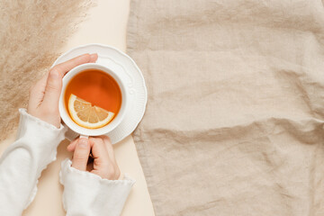 Female hands hold cup of tea with lemon on linen beige cloth background, copy space. Woman with cup...