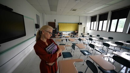 Wide angle view of sad unhappy teacher in large empty classroom with the lights turned off during budget cuts or a pandemic wondering about the future of education.