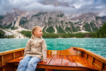 Fototapeta premium Little girl sitting in big brown boat at Lago di Braies lake in cloudy day, Italy. Summer vacation in Europe