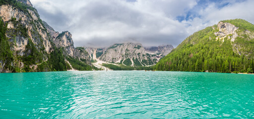 Braies lake surrounded by pine forests and the rocky ranges of the Dolomites in cloudy day, Italy.