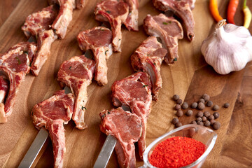 Metal skewers with raw uncooked lamb ribs meat for frying on the wooden board with chili peppers, garlic and spices near it. Close-up perspective view, shallow depth of field. Meat and spices in focus