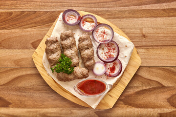 Fried meat kebabs with red onion rings, sauce and pita bread on the wooden board on the wooden table, flat lay top view