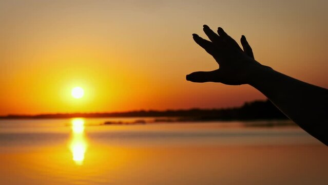 Silhouette hand of a young girl stretches out to the sunset, slow motion. Setting sun rays between fingers woman hand. Palm on the sun, faith in god. Concept dream, happiness, freedom, mental wellness