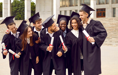 Group of happy successful multiracial students celebrating university graduation day together. Students in academic gowns laughing standing near university stairs. Education and graduation concept.