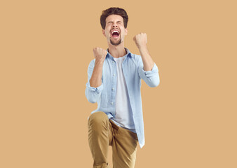 Photo of overjoyed young man screaming happily raising fists. Photo of excited casually dressed man shouting yeah with closed eyes over isolated beige background. Positive human emotions concept