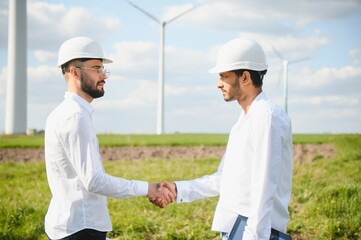 Close up of indian technician and inspector shaking hands while standing on farm with wind turbines. Concept of people, teamwork and eco friendly energy