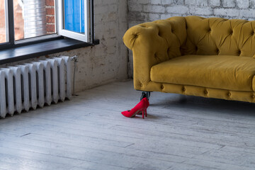 Side view of red stiletto high heel women's shoes standing on wooden floor next to yellow sofa in empty room. No people. Soft focus. Copy space for your text. Footwear theme.