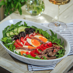 nicoise salad layered in a deep plate served with a glass of wine on a rustic wooden table on a napkin