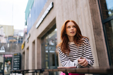 Low-angle view portrait of red-haired attractive young woman sitting at table with smartphone in outdoor cafe terrace in sunny summer day. Happy pretty redhead female lady waiting for someone in cafe.