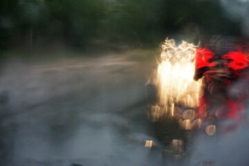 Abstraktes Motiv mit fahrendem Auto, roten und weißen Lichtern auf nasser Straße mit Spiegelung von Lichtern vor grünen Bäumen bei regen und Sturm am Nachmittag im Herbst