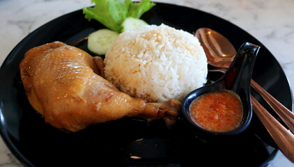 Close up of Chicken braised in soy sauce and rice set on black dish on table with spoon and fork, tangy and spicy sauce in small cup, Thailand.