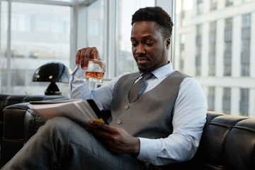 Portrait of successful black businessman drinking whiskey in office and reading documents on leather couch