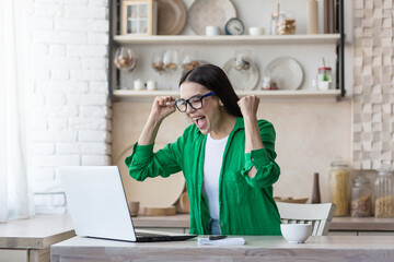 Portrait of a young happy female student sitting at home in the kitchen using a laptop and...