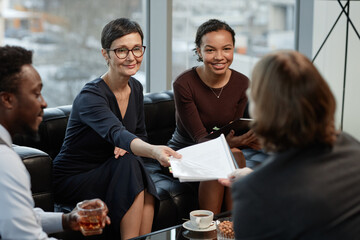 Portrait of smiling mature businesswoman handing documents to colleagues or partners during meeting...