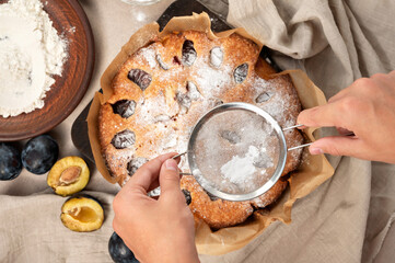 Plum torte decoration with icing. Hands holding sieve with sugar powder above plum pie. Making...