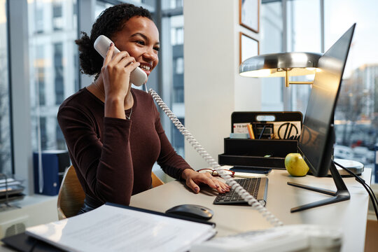 Side view portrait of black young businesswoman answering phone calls at desk in office and smiling - Powered by Adobe