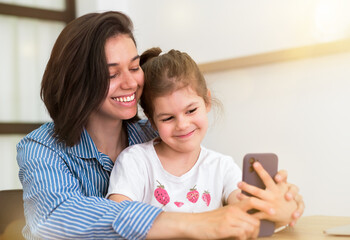 Happy family, cute little kid, daughter with mom, laughing, using smart phone, watching funny video on social media app, taking selfie, looking at mobile phone screen at home