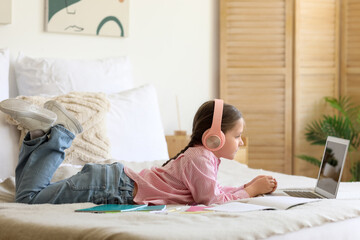 Little girl in headphones with laptop studying computer sciences online on bed at home