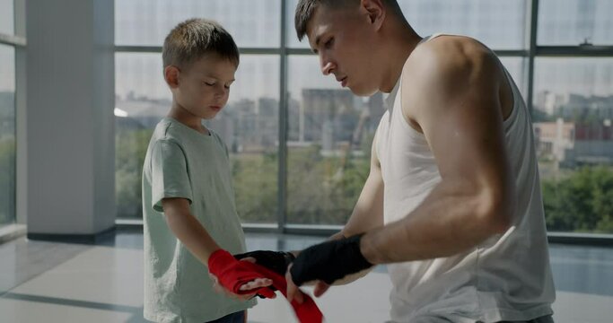 Father and little son boxers preparing for training wrapping hands for protection indoors in gym. Active lifestyle and sports equipment concept.