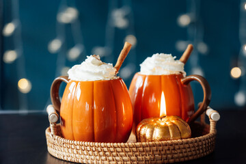 Pumpkin shaped cups of hot drink with whipped cream, cinnamon stick on black table with dark...
