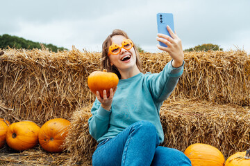 Smiling woman in fun glasses sitting on straw bales and holding pumpkin and making selfie on phone....