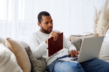 Young african american man in wireless earphone holding notebook and using laptop during online...