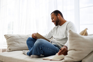 Fototapeta premium Smiling african american man in earphone reading book near notebook on couch at home, self-improvement concept