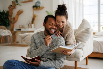 Joyful multiracial couple in warm clothes reading book together while spending time at home