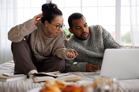 multiracial couple in warm clothes talking near laptop and books during online education at home on bed