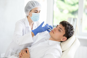 Closeup face of young male client receiving injections during lip enhancement procedure, professional cosmetologist hands in rubber gloves holding syringe