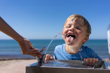 blond boy 3 years old drinks water from a water fountain on the beach in summer