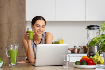 Sporty young woman with healthy smoothie and laptop in kitchen