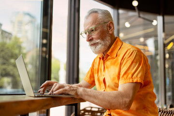 Handsome senior man, programmer using laptop computer working online sitting in modern cafe....