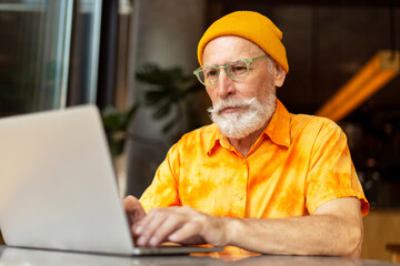 Handsome senior man, bearded programmer wearing yellow hat using laptop computer working online at workplace. Stylish freelancer, copywriter typing on keyboard sitting in modern cafe. Remote job