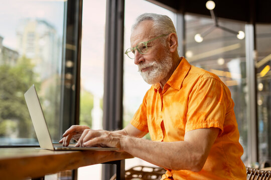 Handsome senior man, programmer using laptop computer working online sitting in modern cafe. Stylish businessman planning startup, searching information on website. Successful business concept