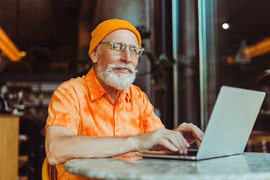 Handsome smiling senior man, bearded hipster wearing yellow hat using laptop computer, working online sitting in modern cafe. Stylish businessman planning startup. Successful business concept  