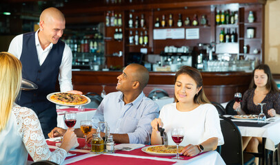 Fototapeta na wymiar Portrait of polite smiling waiter serving pizza to friendly company in cozy pizzeria