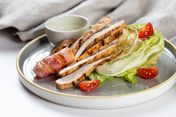 Caesar salad with chicken slices, iceberg lettuce, cherry tomatoes, bacon and sauce on a gray plate on a white background with cloth