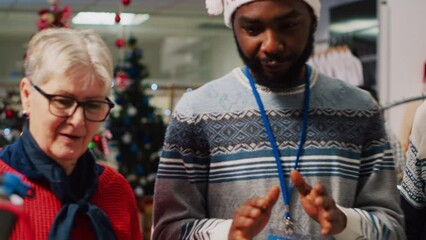 Retail assistant in festive decorated clothing store helping elderly couple during their Christmas frenzy shopping spree. Employee offering advice to senior customers during winter holiday season
