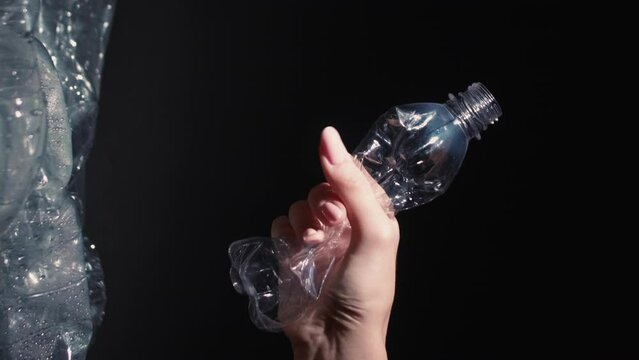 Vertical video. Waste reduction. Plastic recycling. Woman volunteer hand crushing used bottle throwing into trash dump with cellophane bags isolated on black background.