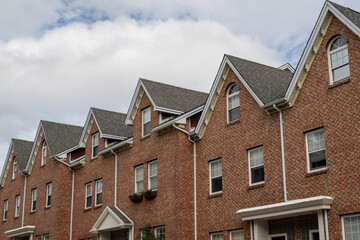 A row of brown brick townhouses with peaked gable roofs, and double hung windows with white trim. The shingles are black asphalt. The adjoined facade row houses are multi-story residential buildings. 