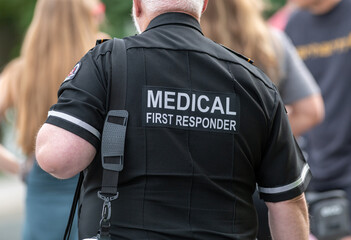 A close-up of a Caucasian male emergency health medical first responder or paramedic wearing a black uniform with grey letters. The ambulance attendant is wearing a short-sleeved shirt with lettering.