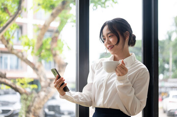 A beautiful Asian businesswoman sipping coffee and using her phone while standing by the window