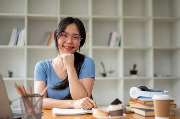 A beautiful Asian female college student in glasses sits at her study table at home.