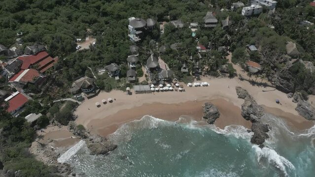 Beach aerial tilts over grass roof resort hotel on rocky sand crescent