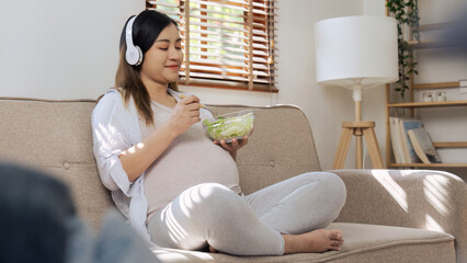 Young pregnant woman listening to music and eating salad on sofa in living room
