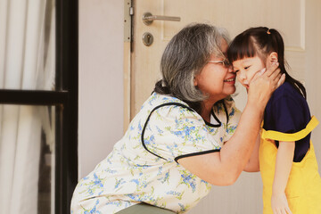 Adorable granddaughter showing love, bonding, warm heart, standing and kissing Grandma's cheek in front of the door before going to kindergarten. - obrazy, fototapety, plakaty