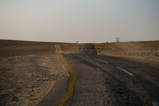 A curving, windy road goes of into the distance, vanishing behind a hill. There is nothing around the road, in the barren, remote wilderness of the Judean Desert in southern Israel.