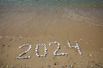 New Year 2024, lettering on the beach with wave and clear blue sea. Numbers 2024 year on the sea shore, message handwritten in the golden sand on beautiful beach background. New Years concept.
